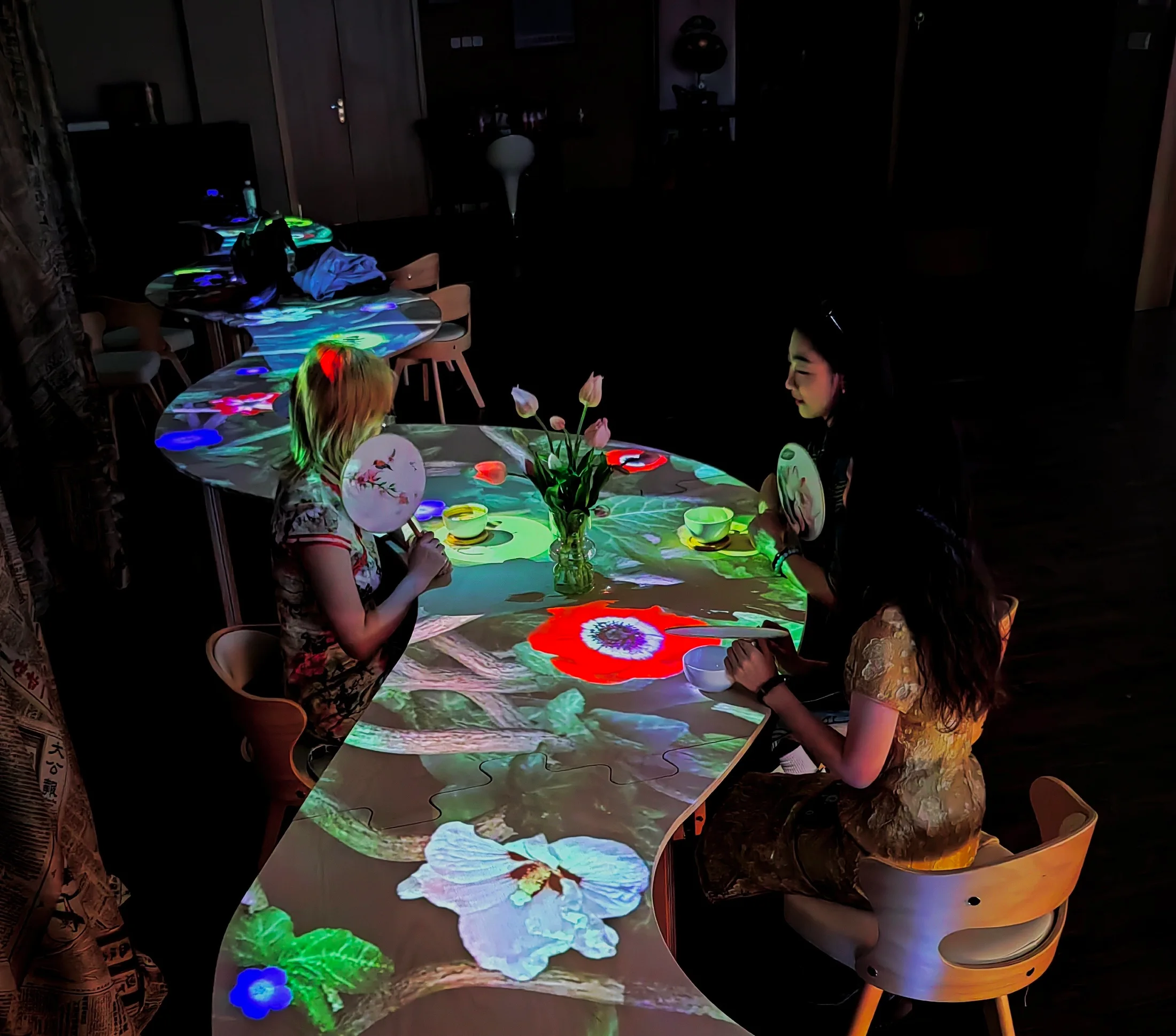 Three visitors around the curved café table, with projected flowers, cups and a vase.