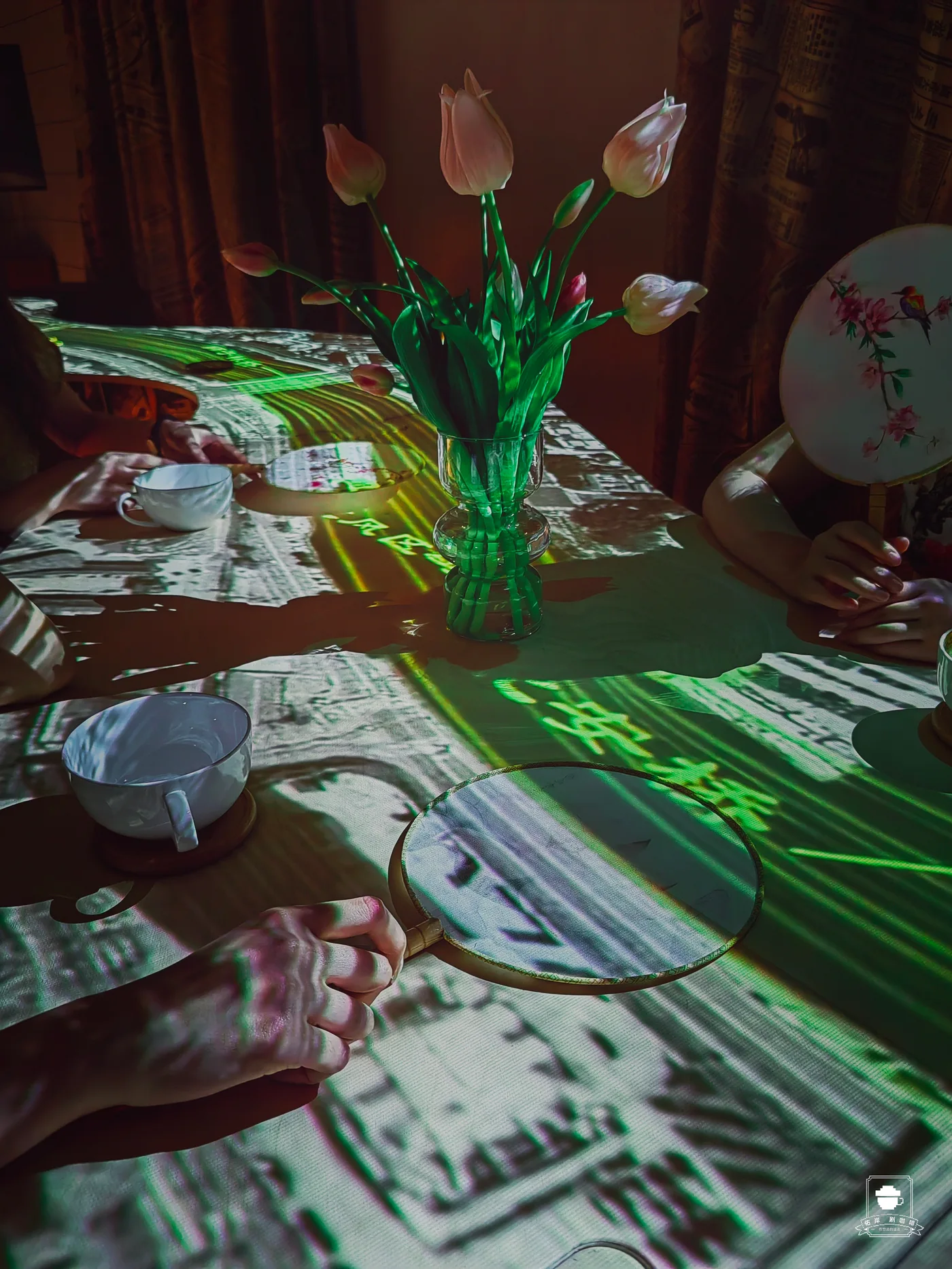 Hands, magnifying glasses, cups and a vase share a tabletop covered in newspaper projection.